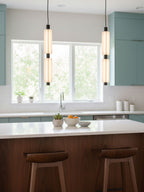 Two-head Clérachic pendants with black joints hang in front of a kitchen window, above a white island with walnut stools.
