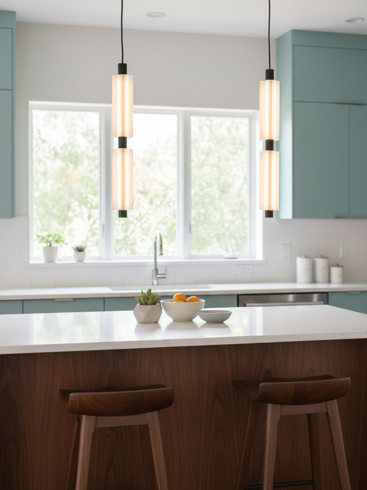 Two-head Clérachic pendants with black joints hang in front of a kitchen window, above a white island with walnut stools.