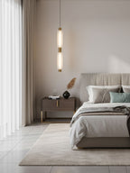 A single two-head Clérachic pendant hangs beside a bed in a pale bedroom with long curtains and a woven rug.