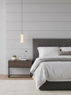 A small single-head Clérachic pendant hangs in front of a panelled wall, beside a grey bed and a dark metal bedside table.