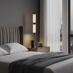 The soft glow of a two-head Clérachic pendant sits above a bedside shelf, with a charcoal throw on the bed below.