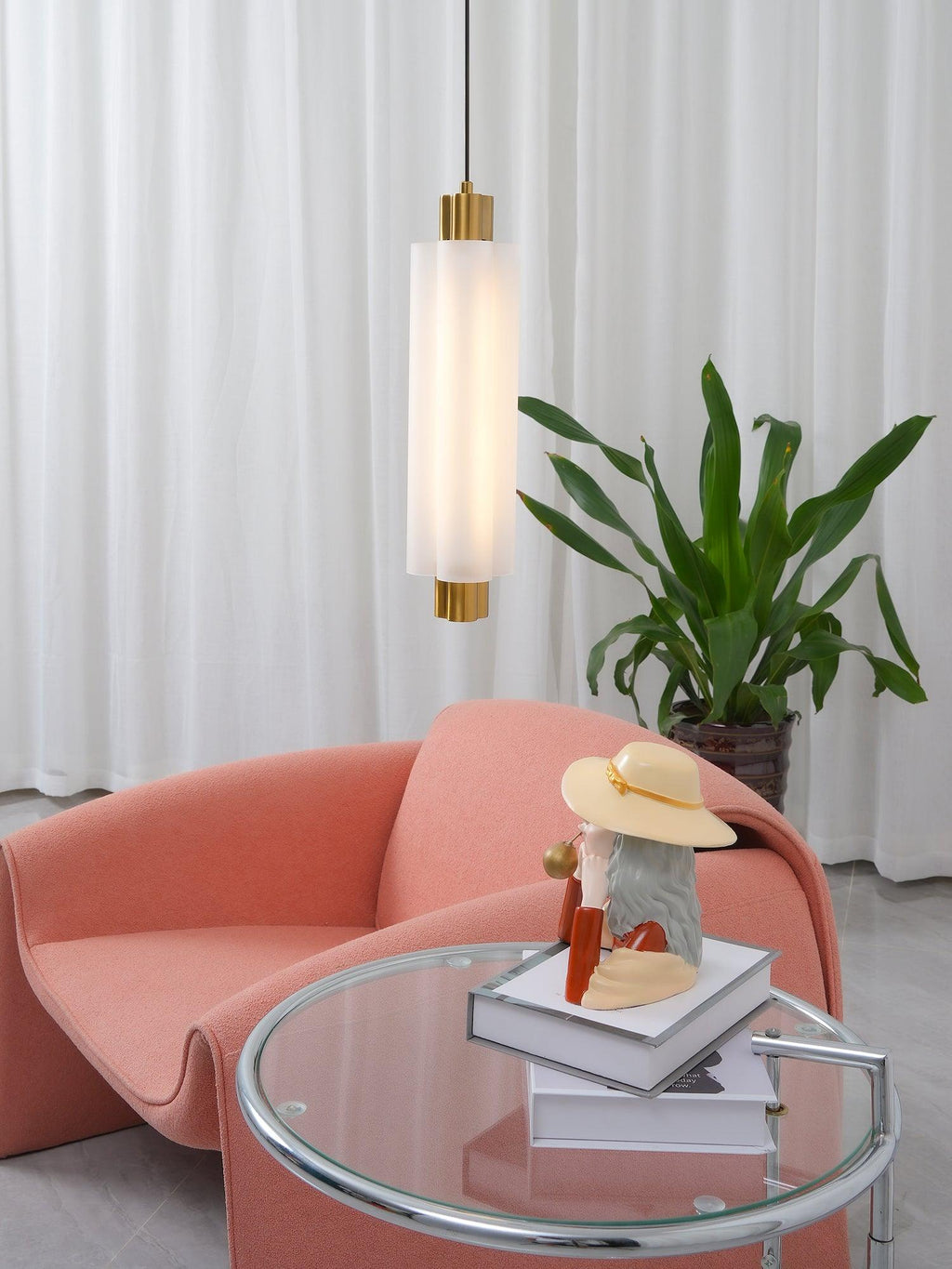 A frosted Clérachic pendant with gold ends hangs above a coral armchair, with a glass side table and a leafy plant.
