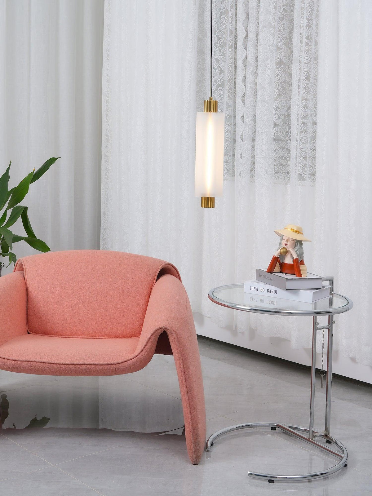 A lit single-head Clérachic pendant with gold ends hangs near sheer curtains, with a coral chair and a glass table below.