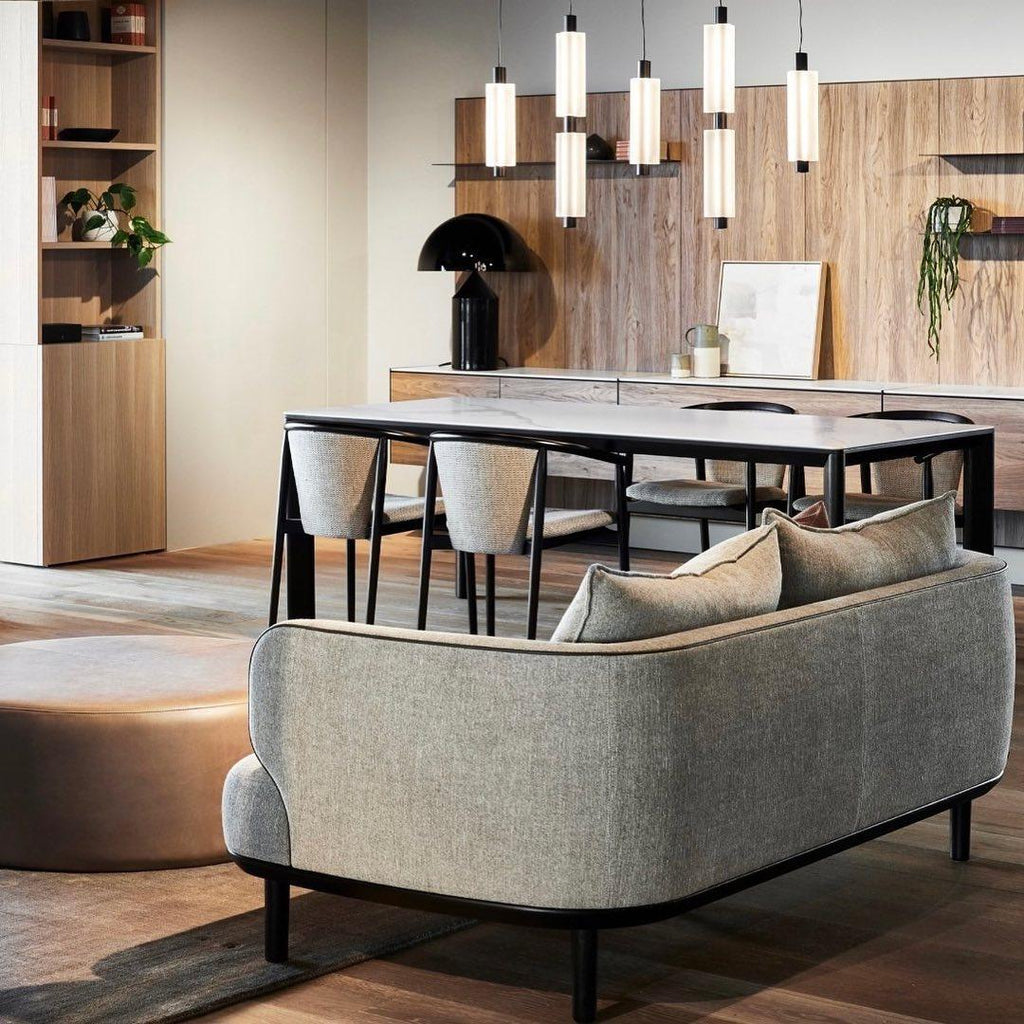 Several Clérachic pendants hang over a dining table, with a grey curved sofa and a round leather pouffe in the foreground.