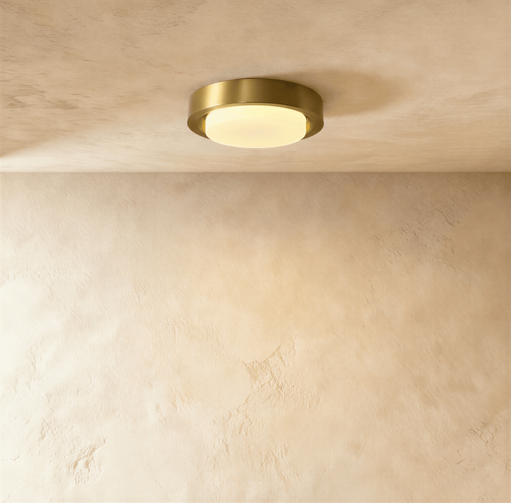 Clérachic round flush ceiling light with a brushed brass rim and glowing frosted glass cover, against a limewashed plaster ceiling.