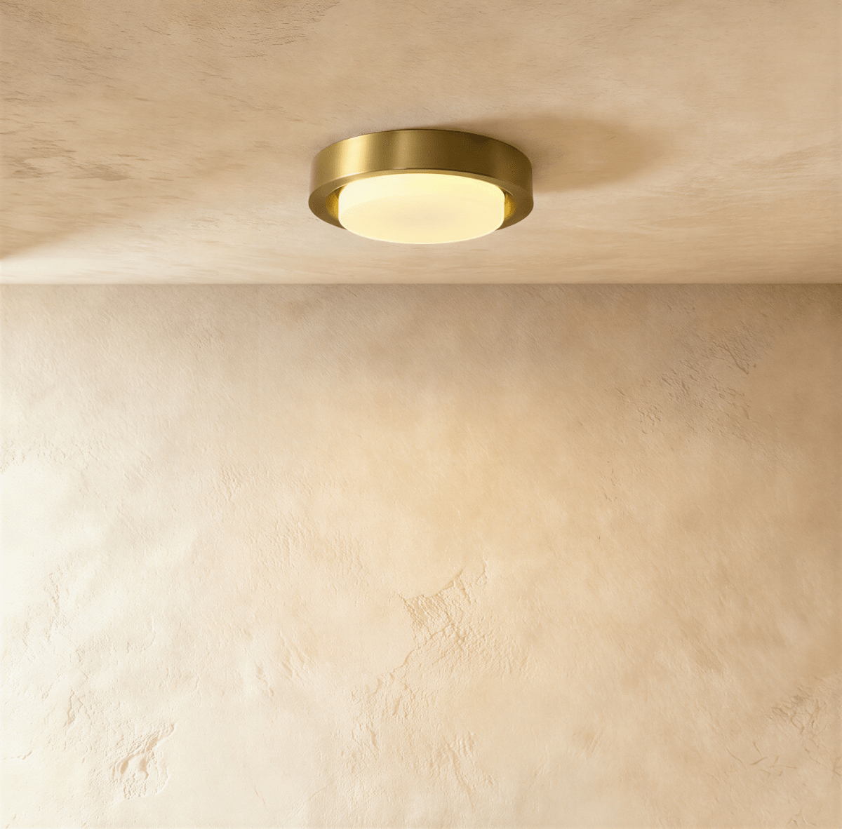 Clérachic round flush ceiling light with a brushed brass rim and glowing frosted glass cover, against a limewashed plaster ceiling.