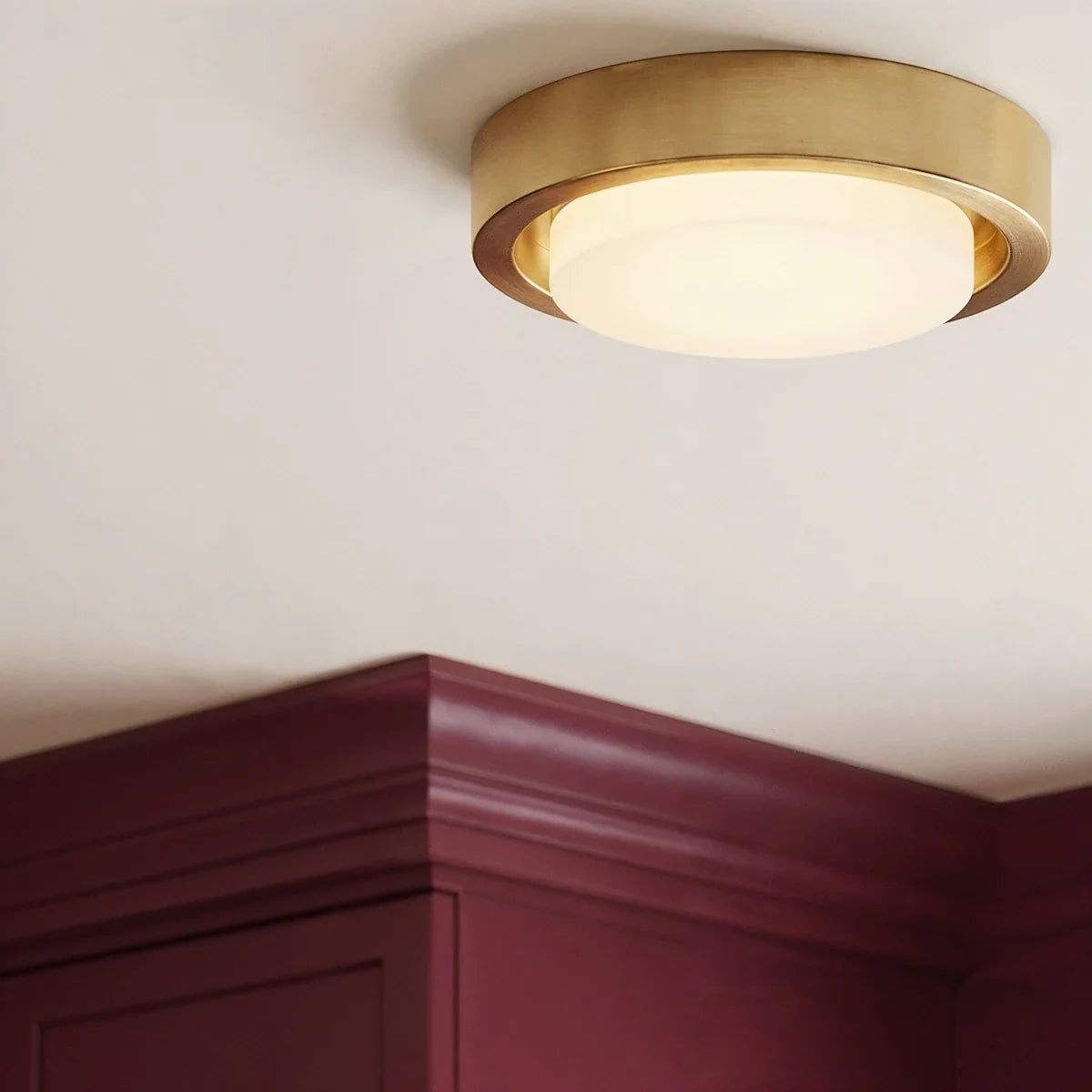 Close view of a Clérachic brass flush ceiling light with a lit frosted glass cover, set above burgundy panelled cornicing.