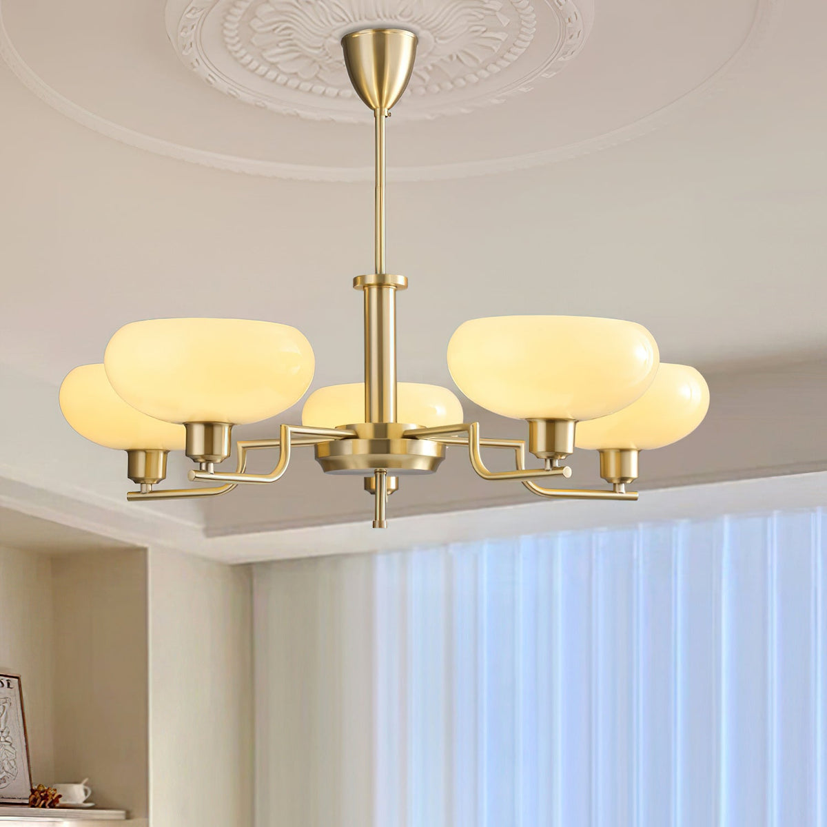Gold Clérachic chandelier lit beneath a plaster ceiling rose, its beige glass heads glowing above a window with sheer curtains.