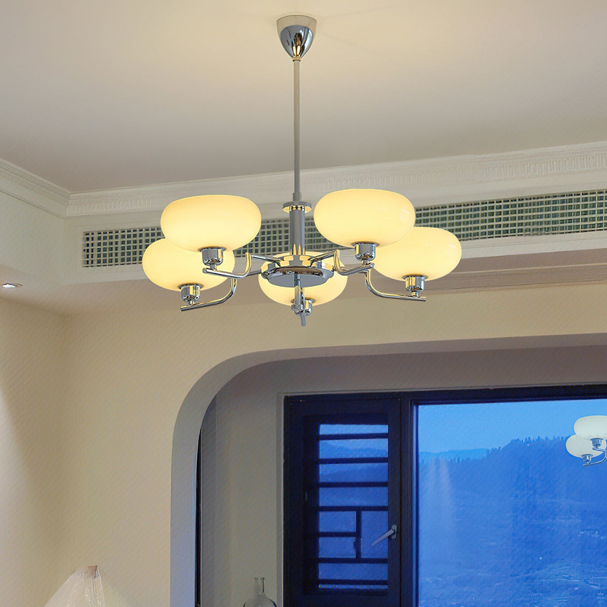 Chrome Clérachic chandelier lit high in a room with a cream ceiling and a ceiling vent, an arched window with a mountain view below.