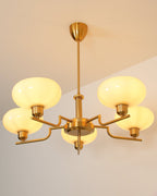 Gold Clérachic chandelier lit against a peach wall and ceiling, five glowing butter-yellow glass heads on brass arms.