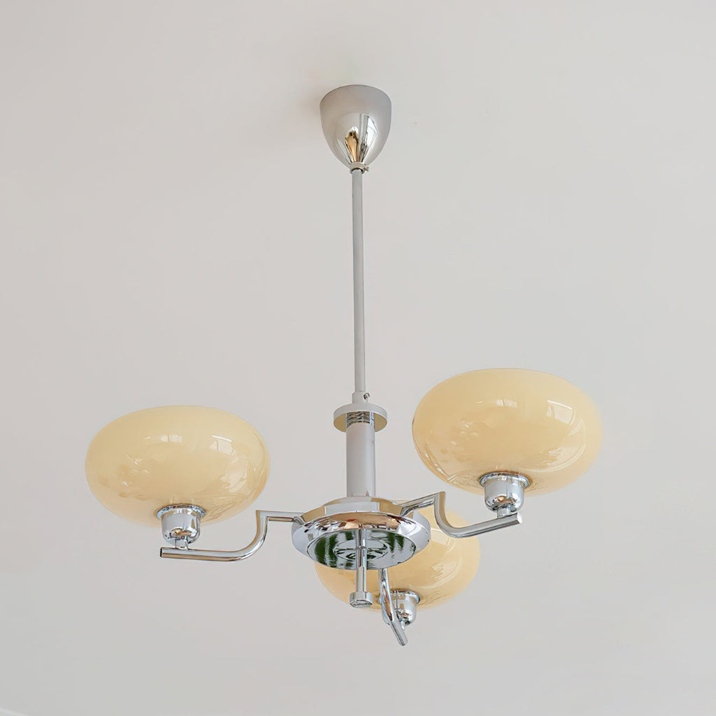 Three-head chrome Clérachic chandelier unlit against a white wall, with beige glass domes on curved polished arms.