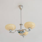 Three-head chrome Clérachic chandelier unlit against a white wall, with beige glass domes on curved polished arms.