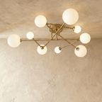 Clérachic nine-head ceiling light with intersecting brass-toned arms and frosted glass globes in two sizes, lit against limewash.
