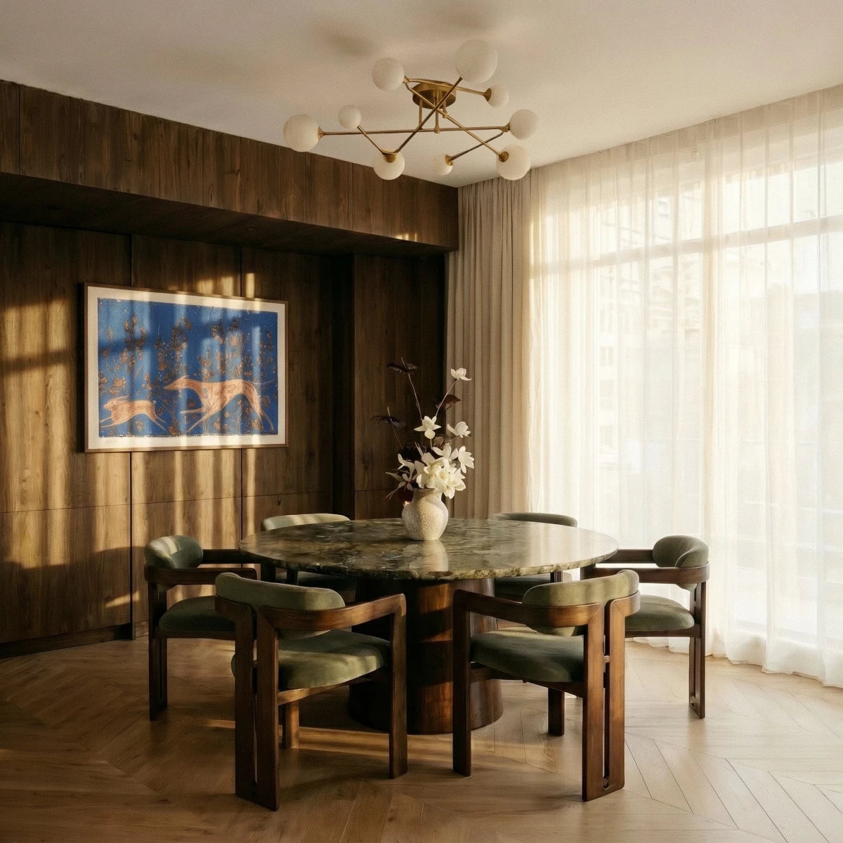 Clérachic nine-head brass-toned ceiling light with frosted globes over a round stone-topped table with green armchairs.