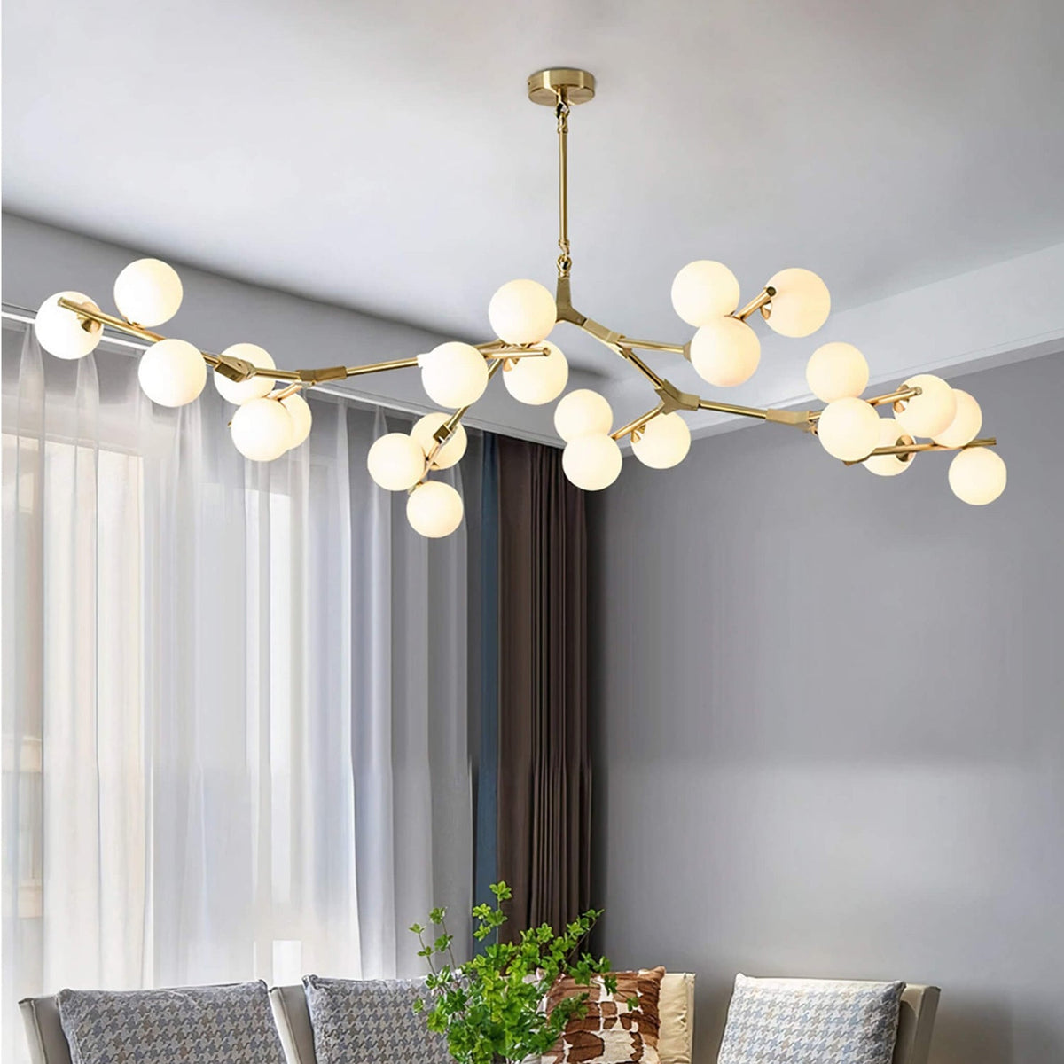 This Clérachic chandelier in gold and white hangs in a grey living room, its branches carrying frosted globes beside sheer curtains.