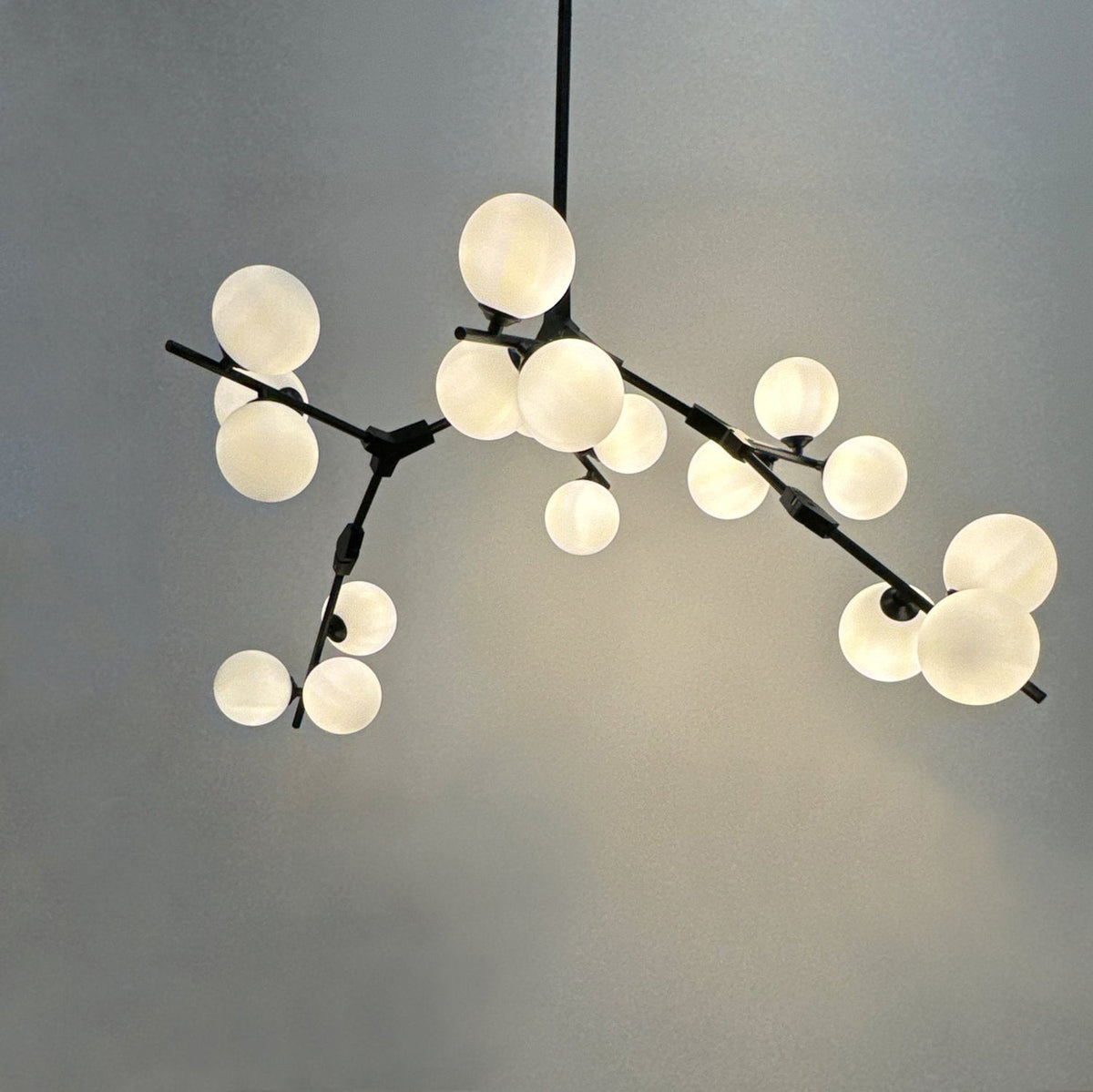 Close view of the lit chandelier, warm frosted globes of different sizes on black branching arms against a pale grey-green wall; Clérachic.