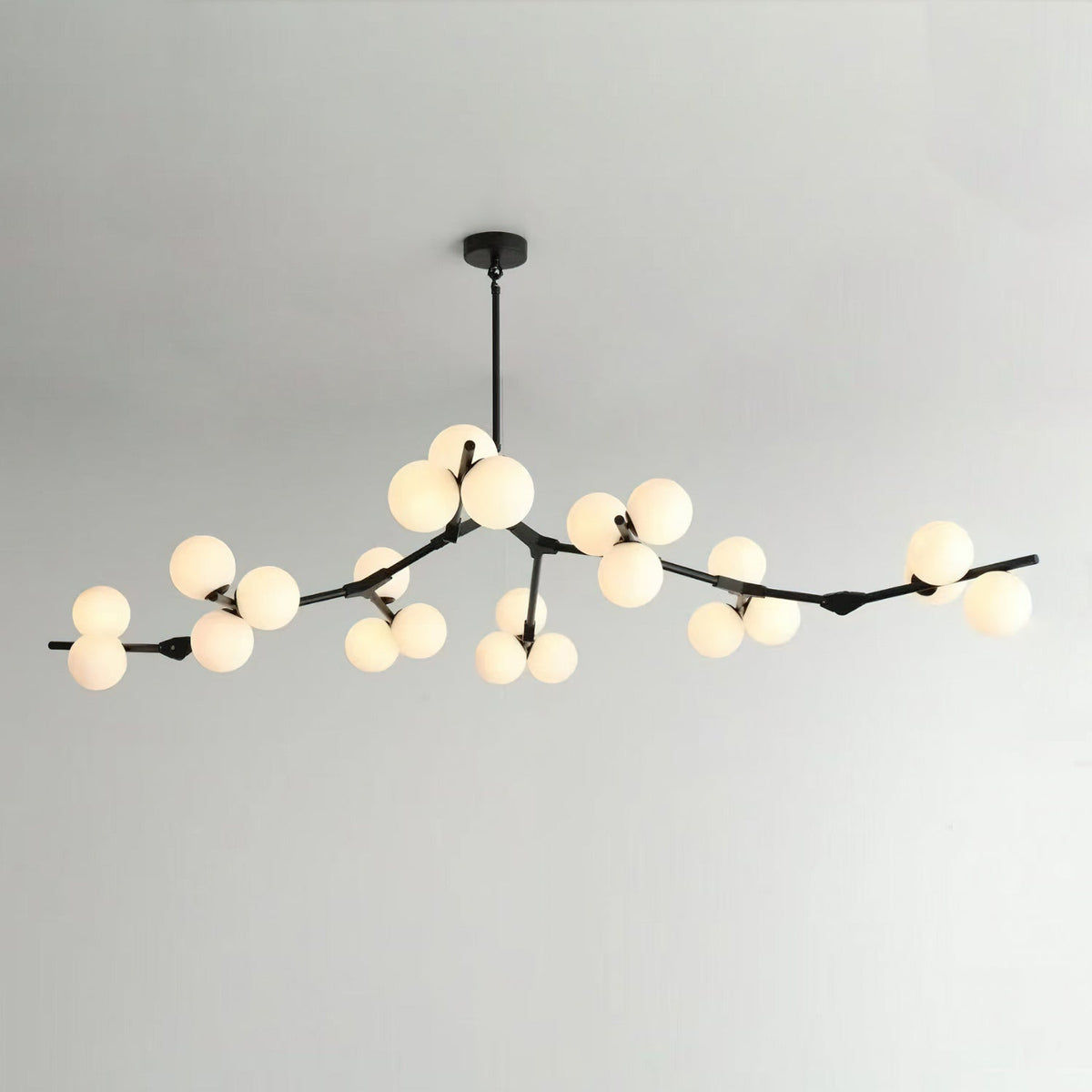 Black Clérachic chandelier on a thin rod with many lit frosted globes along long, low branches, against a pale grey wall.