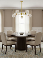 White globes on a gold Clérachic chandelier above a round dark dining table with cream upholstered chairs and a window with curtains.