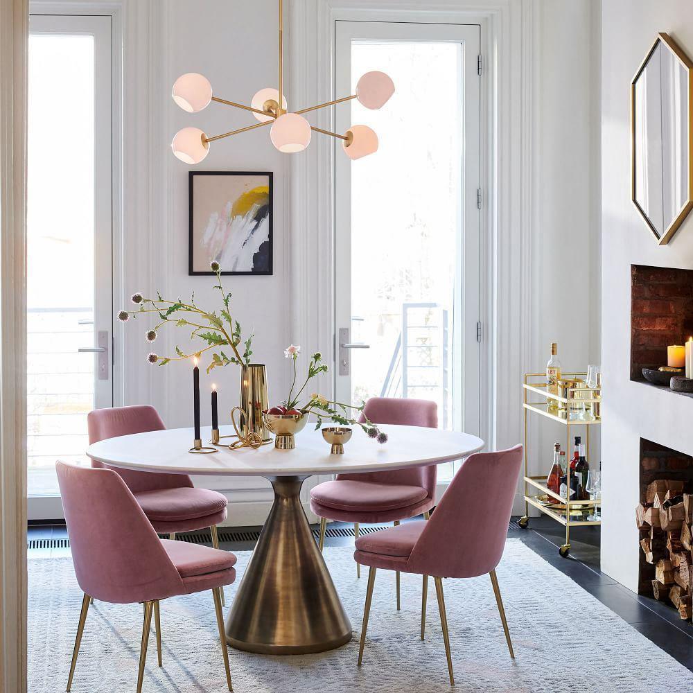 Gold chandelier from Clérachic with blush-lit white globes above a round dining table with pink chairs, a brass-based table and a fireplace.