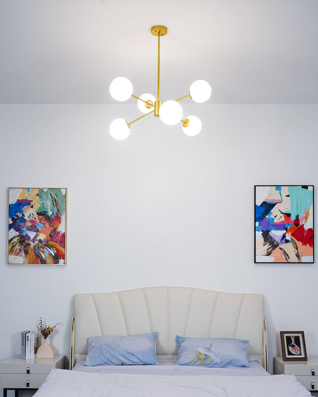 Small gold chandelier from Clérachic with white globes lit above a bed with a cream headboard and two colourful framed prints.