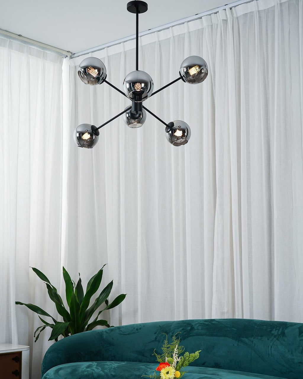 Black chandelier from Clérachic with six smoked glass globes in front of white curtains, with a plant and a green velvet sofa below.