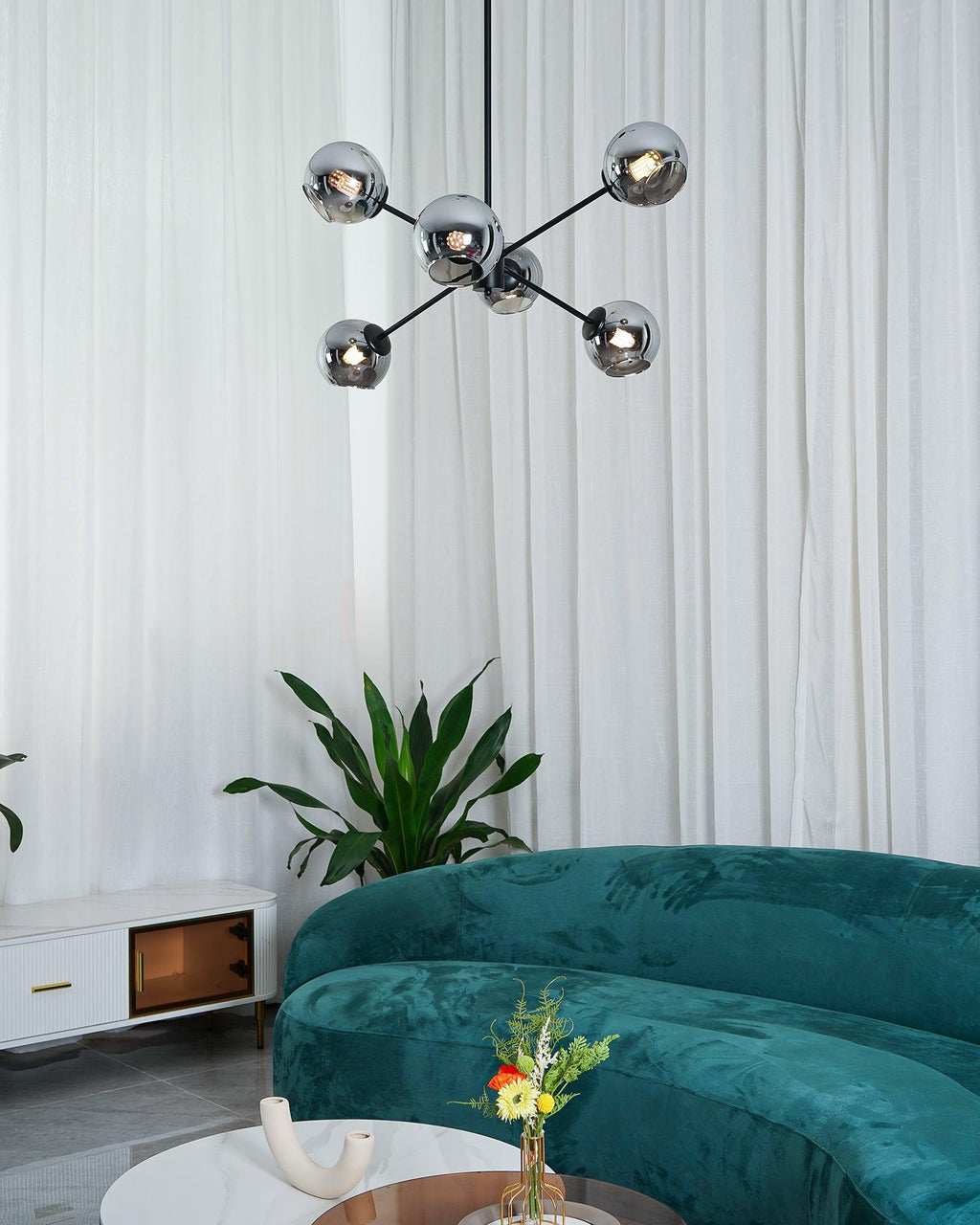 Black Clérachic chandelier with smoked glass globes, seen from a distance with white curtains, a plant and a green sofa below.