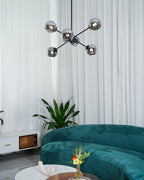 Black Clérachic chandelier with smoked glass globes, seen from a distance with white curtains, a plant and a green sofa below.