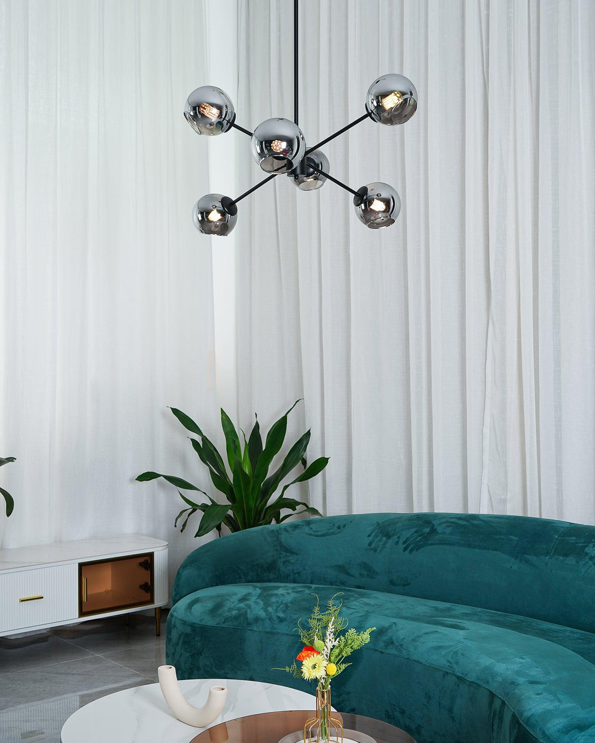 Black Clérachic chandelier with smoked glass globes, seen from a distance with white curtains, a plant and a green sofa below.