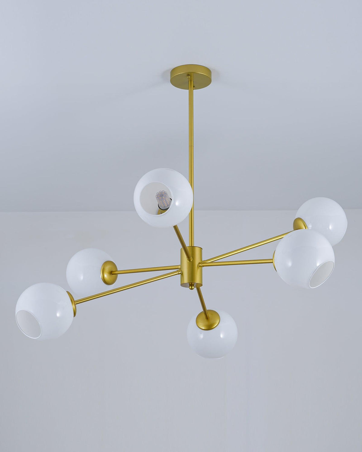 Gold chandelier from Clérachic with white globes and open-sided shades, hung from a slim rod below a plain ceiling.