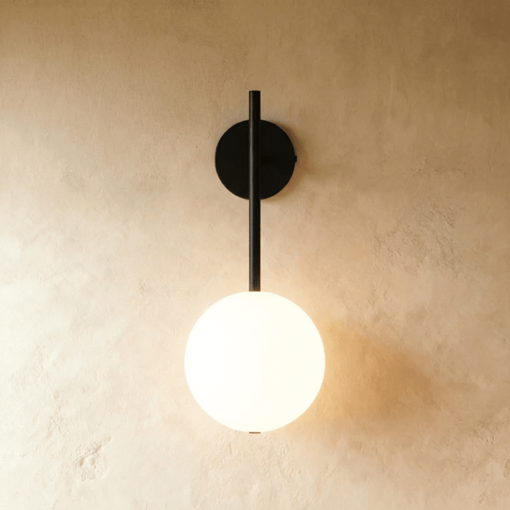 Clérachic wall lamp in black, a slim rod dropping from a round wall plate to a lit frosted glass sphere that casts a warm glow across bare plaster.