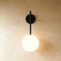 Clérachic wall lamp in black, a slim rod dropping from a round wall plate to a lit frosted glass sphere that casts a warm glow across bare plaster.