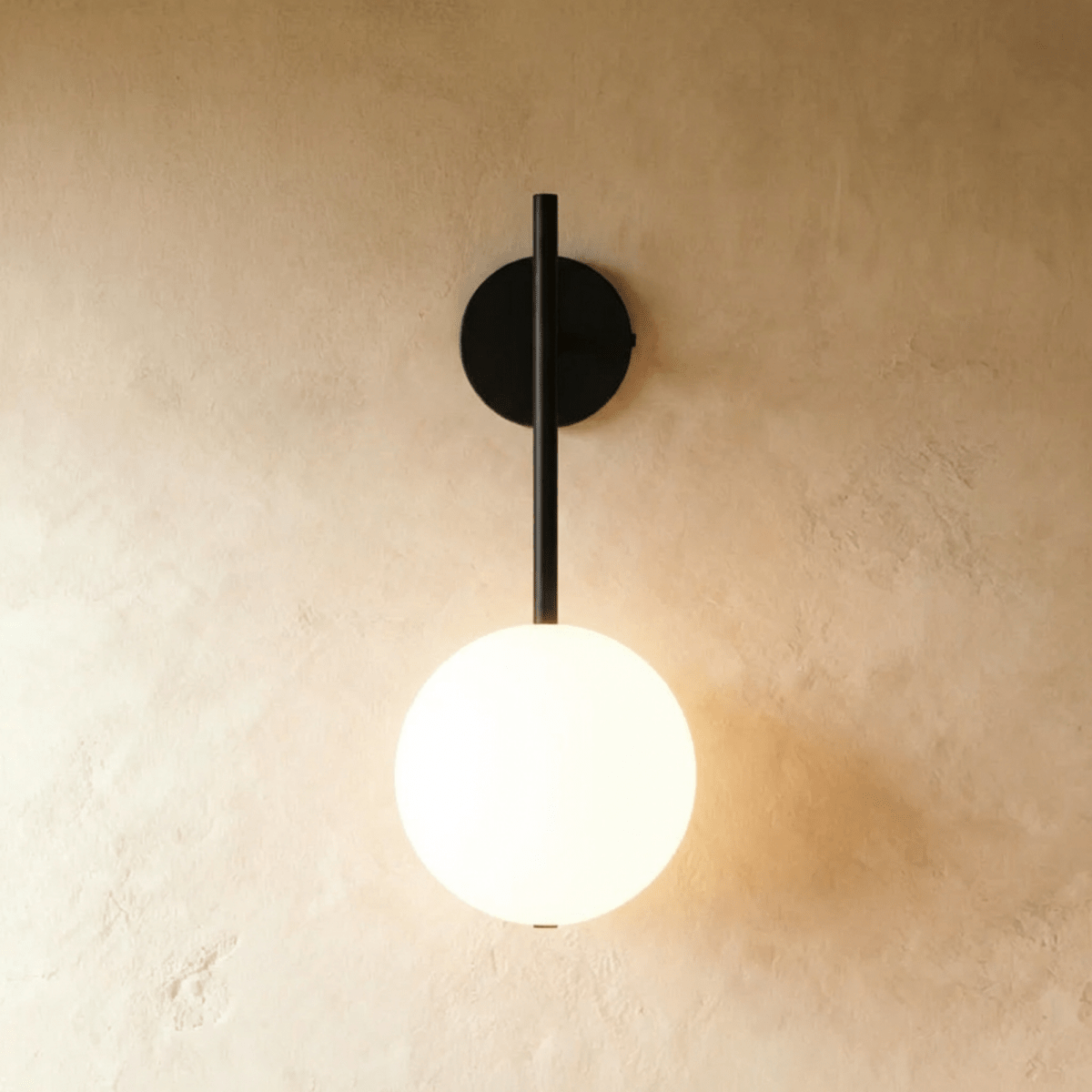 Clérachic wall lamp in black, a slim rod dropping from a round wall plate to a lit frosted glass sphere that casts a warm glow across bare plaster.