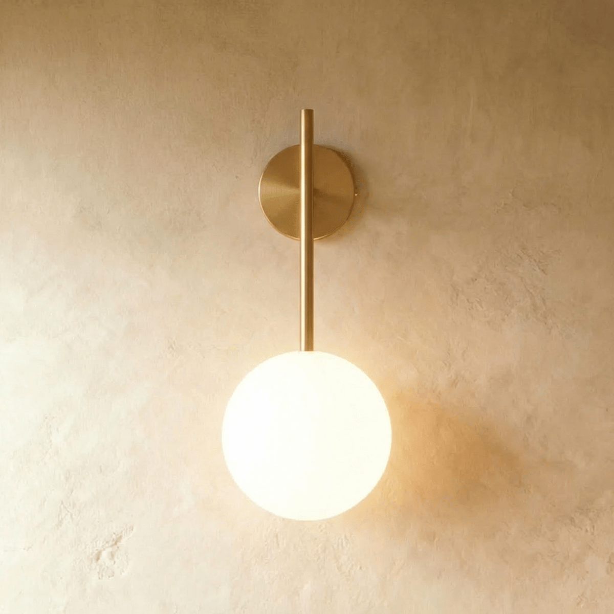 Clérachic wall lamp in warm gold, the same straight rod and lit glass sphere throwing a soft pool of light onto the plaster behind it.
