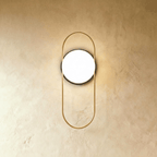 Clérachic wall lamp with a tall gold loop standing proud of a round black backplate, the lit glass sphere held in the upper half of the loop.
