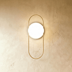 Clérachic wall lamp with a tall gold loop and a round white backplate, the lit glass sphere sitting high inside the loop.