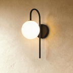 Clérachic wall lamp in black, a hooked arm curving out from a round wall plate to carry the lit sphere clear to one side.