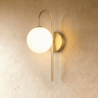 Clérachic wall lamp in gold, the hooked arm holding the lit sphere away from its round wall plate against warm plaster.