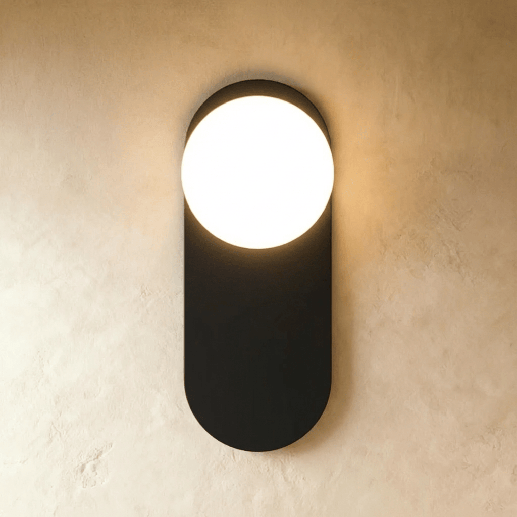 Clérachic wall lamp in black, a flat capsule-shaped plate with the lit sphere breaking out of its top edge and light spilling above and below.