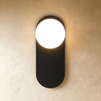 Clérachic wall lamp in black, a flat capsule-shaped plate with the lit sphere breaking out of its top edge and light spilling above and below.