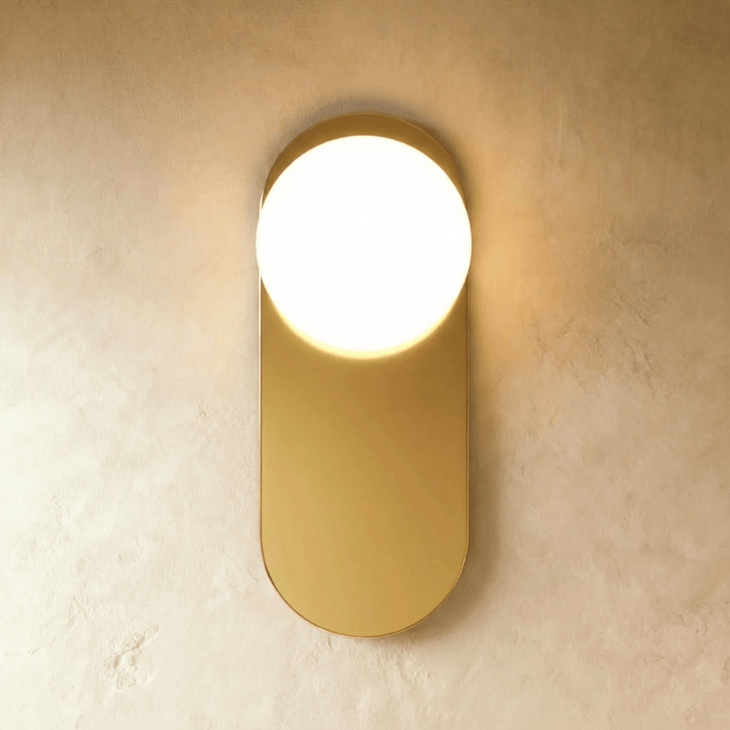 Clérachic wall lamp in gold, the same flat capsule plate lit so the sphere reads bright against the brass face.