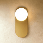 Clérachic wall lamp in gold, the same flat capsule plate lit so the sphere reads bright against the brass face.