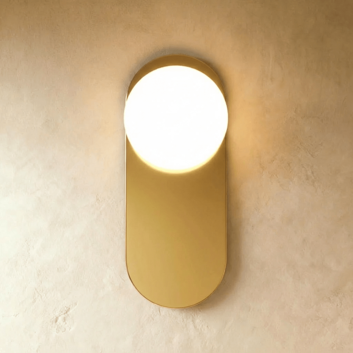 Clérachic wall lamp in gold, the same flat capsule plate lit so the sphere reads bright against the brass face.