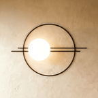 Clérachic wall lamp in black, a wide ring crossed by two parallel rods that run well past it on both sides, the lit sphere set left of centre.