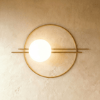 Clérachic wall lamp in gold, the wide ring and paired rods with the lit sphere placed about a third of the way in from the left.