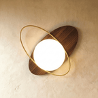 Clérachic wall lamp with a walnut-finished elliptical disc showing its wood grain, tilted and threaded by a gold ring around the lit sphere.