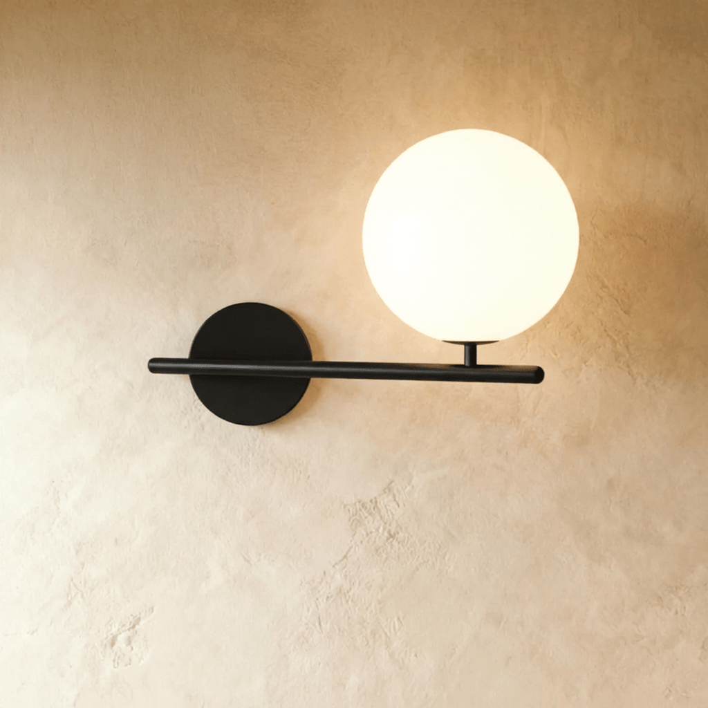 Clérachic wall lamp in black, a horizontal bar crossing the wall past its round plate on one side and past the lit sphere on the other, the sphere raised on a short stem.
