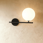 Clérachic wall lamp in black, a horizontal bar crossing the wall past its round plate on one side and past the lit sphere on the other, the sphere raised on a short stem.