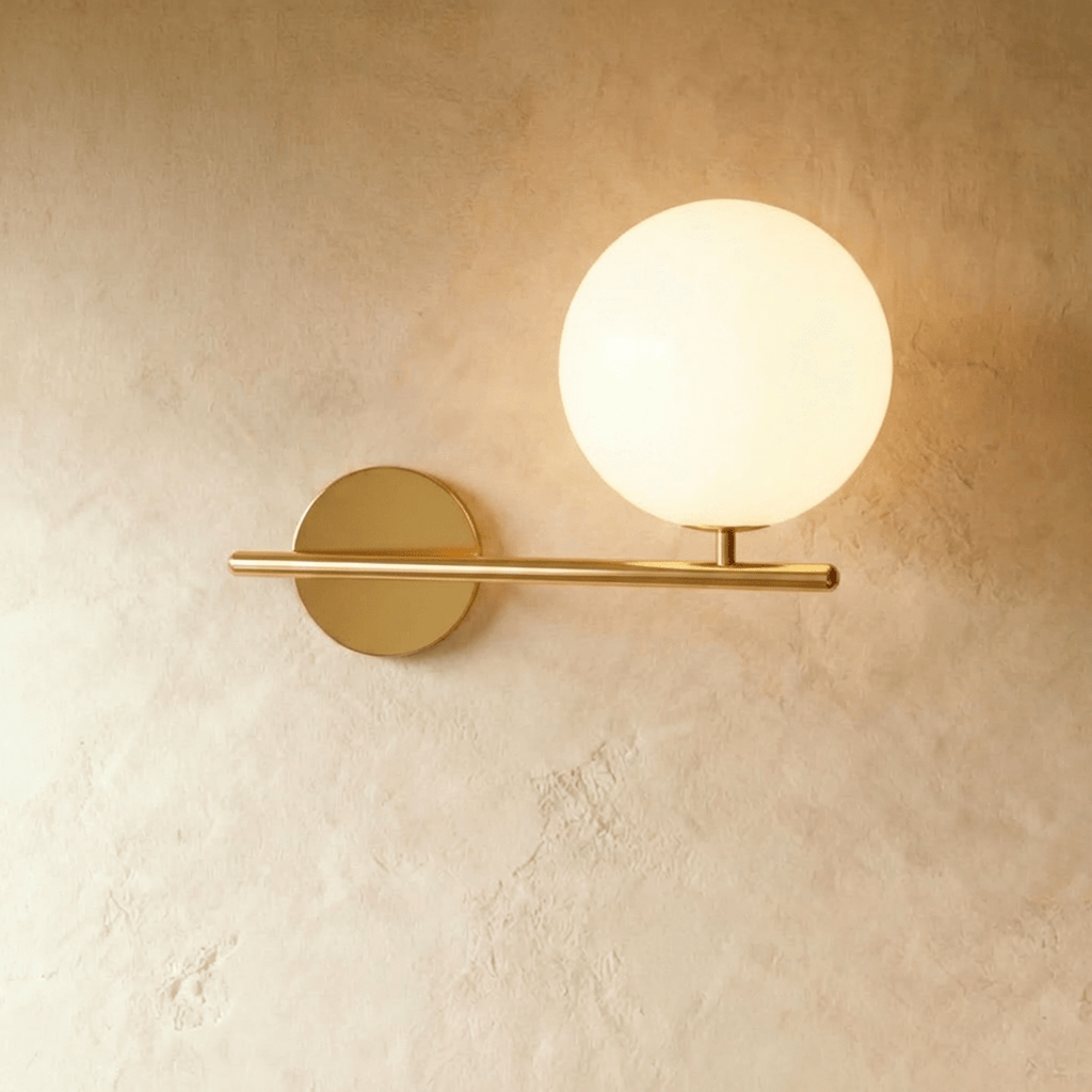Clérachic wall lamp in gold, the same horizontal bar and lit sphere on its short stem, the brass warm against plaster.