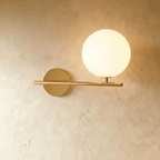Clérachic wall lamp in gold, the same horizontal bar and lit sphere on its short stem, the brass warm against plaster.