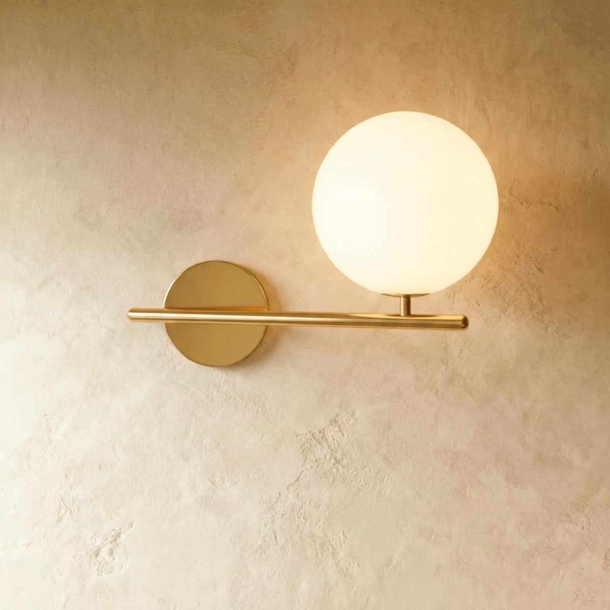 Clérachic wall lamp in gold, the same horizontal bar and lit sphere on its short stem, the brass warm against plaster.